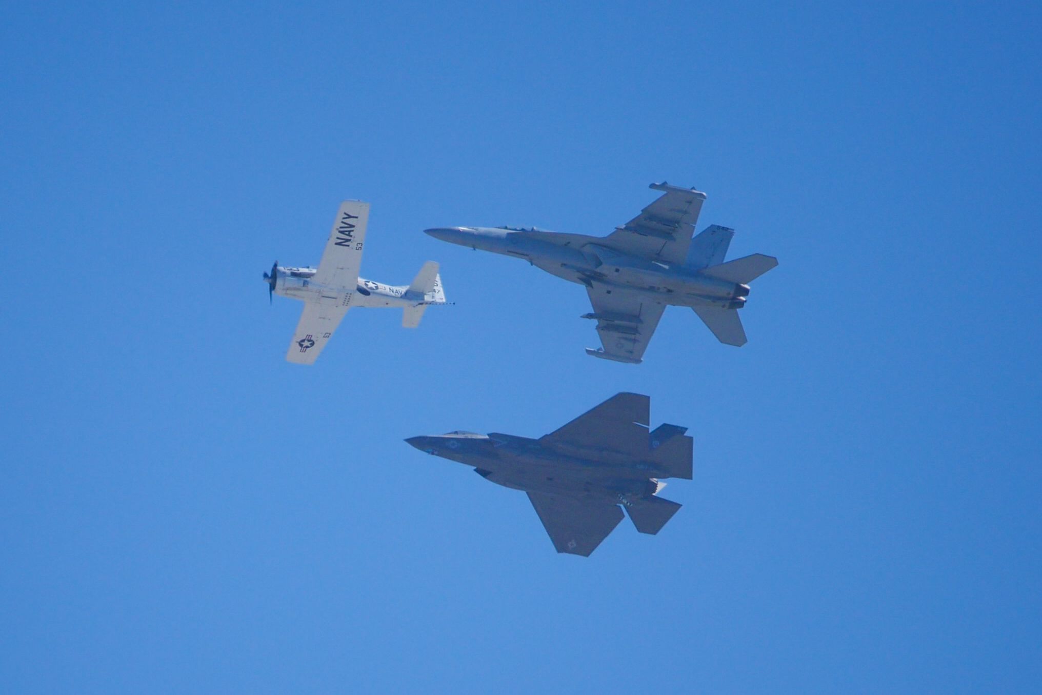 Three planes fly together in formation. An old white prop plane leads as two modern military jets follow.